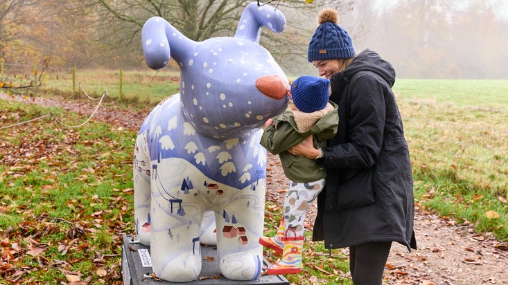 Woman bobble hat lifting up a toddler to touch the nose of a large snow dog sculpture, which is decorated with an alpine ski view.